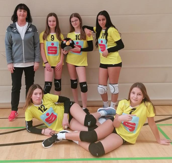 Mädchenvolleyballmannschaft steht mit Trainerin vor einer weißen Wand. Ein Mädchen hält einen Ball in der Hand.